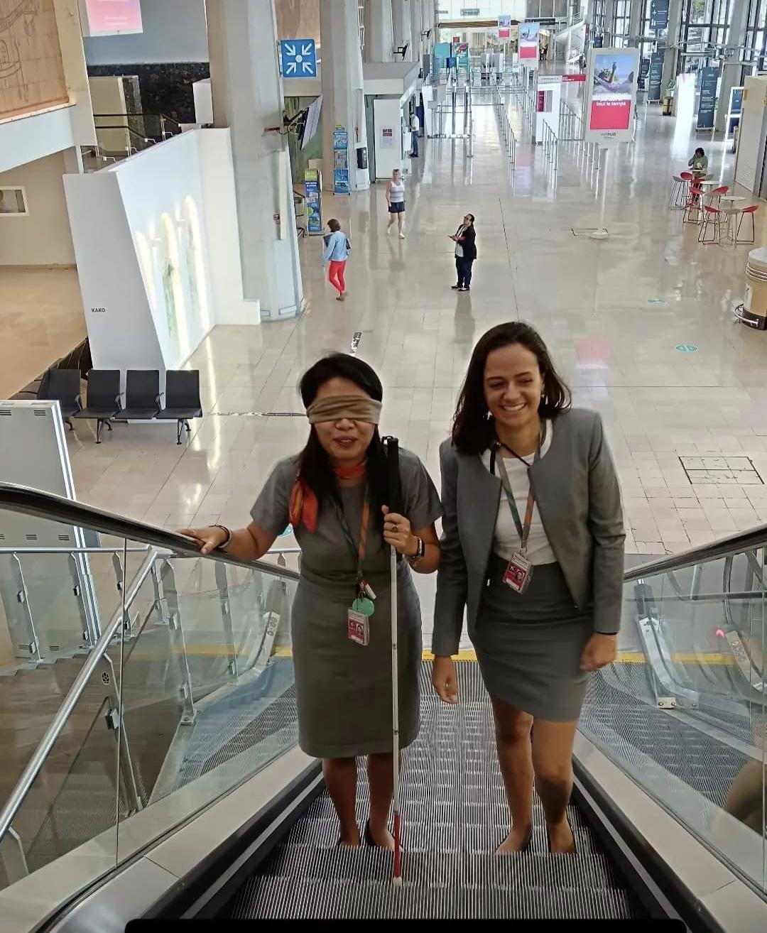 Li et Charlotte (équipe accueil FRT) sur un escalator. Li a les yeux bandés et une canne pour mieux comprendre le déplacement lorsqu'on est porteur d'un handicap visuel