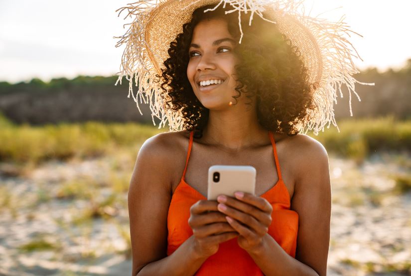 Réunionnaise avec un chapeau de paille sur la plage qui sourit, en tenant son smartphone dans les mains