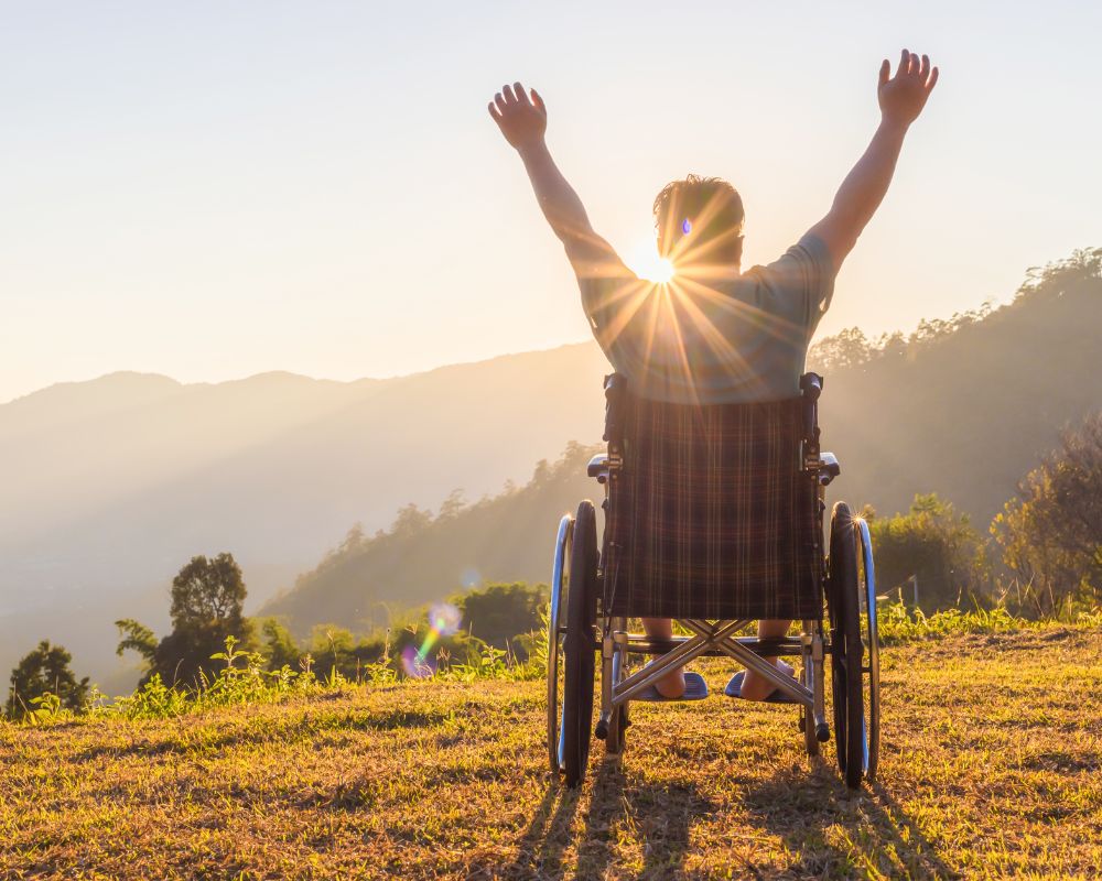 Personne en fauteuil roulant, de dos, devant un coucher de soleil qui lève les bras de façon joyeuse